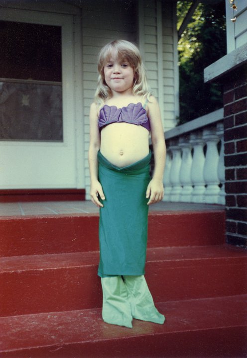 A white child with long blond hair stands proudly on red stairs outside a white house, wearing a handmade mermaid costume.